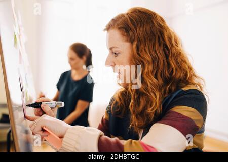 Side view of confident redhead businesswoman writing on whiteboard while female colleague working at creative office Stock Photo