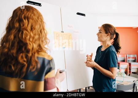 Confident female colleagues discussing plastic management project over whiteboard at creative office Stock Photo