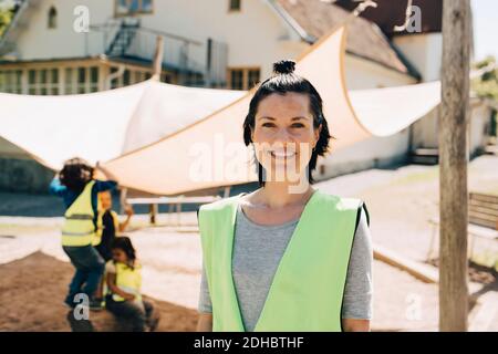 Portrait of confident female teacher standing in reflective clothing against students playing at background Stock Photo