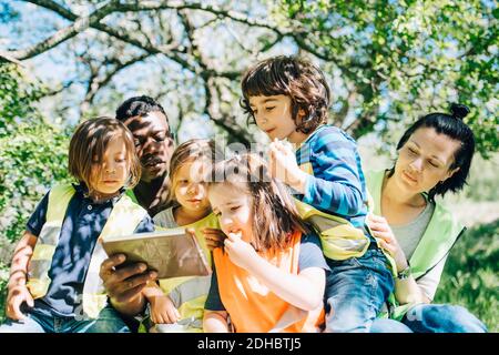 Multi-ethnic students and teachers sharing digital tablet while sitting against trees in playground Stock Photo