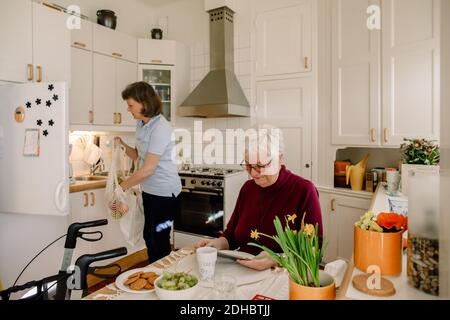 Senior woman using digital tablet while female healthcare worker holding grocery bag in kitchen Stock Photo