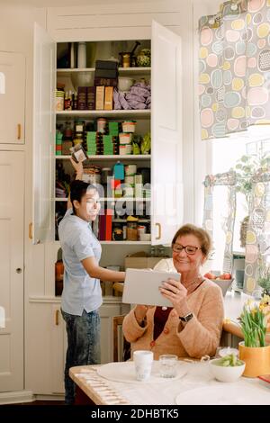 Smiling elderly woman showing digital tablet to young female caregiver working in kitchen at nursing home Stock Photo
