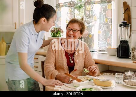 Cheerful senior woman talking with female caregiver in kitchen at nursing home Stock Photo