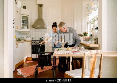 Young female volunteer assisting retired elderly man with rollator in kitchen at nursing home Stock Photo