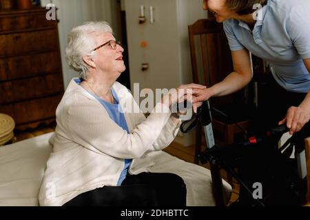 Smiling senior woman holding hands of female healthcare worker in bedroom at nursing home Stock Photo