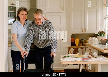 Mature female caregiver assisting retired man walking with rollator in kitchen at nursing home Stock Photo