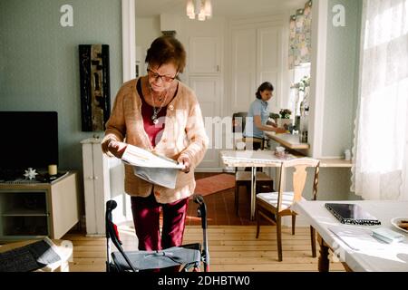 Senior woman reading newspaper while caregiver working in kitchen at nursing home Stock Photo