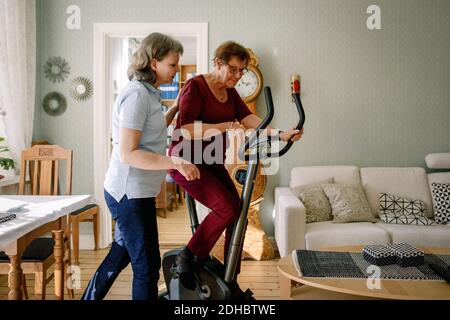 Mature female physical therapist assisting elderly woman on exercise bike at nursing home Stock Photo