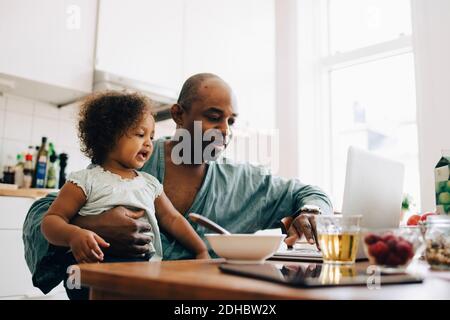 Father carrying daughter while using laptop during breakfast at table in dining room Stock Photo