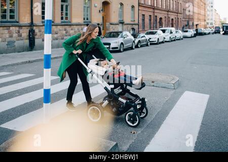 Woman pushing daughter with digital tablet in carriage while crossing city street Stock Photo