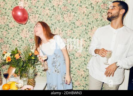 Young man looking at woman playing with balloon while standing against wallpaper at home during dinner party Stock Photo