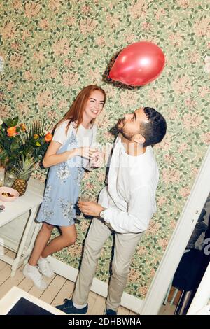 Tilt shot of young man playing with balloon while standing by woman against wallpaper during dinner party at home Stock Photo