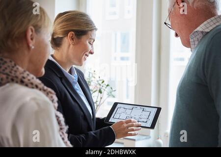Female real estate agent showing new house blueprint on digital tablet to senior couple at home Stock Photo