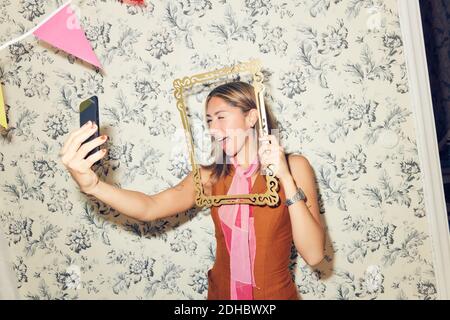 Cheerful young woman taking selfie through picture frame while winking at smart phone against wallpaper during party in Stock Photo