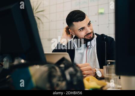 Confused young male computer programmer scratching head while sitting with laptop at creative office Stock Photo