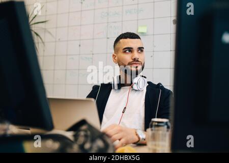 Young male computer hacker sitting with laptop while looking away at creative office Stock Photo