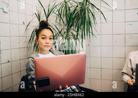 Portrait of confident young female hacker using laptop while sitting against tile wall at small creative office Stock Photo