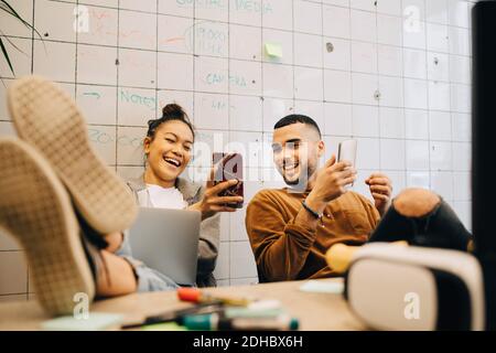 Smiling young businesswoman sitting with feet up on desk sharing smart phone with businessman against wall at creative o Stock Photo