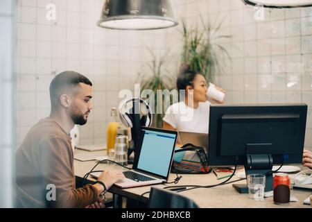 Young male hacker using laptop while female colleague sitting in creative office seen through window Stock Photo