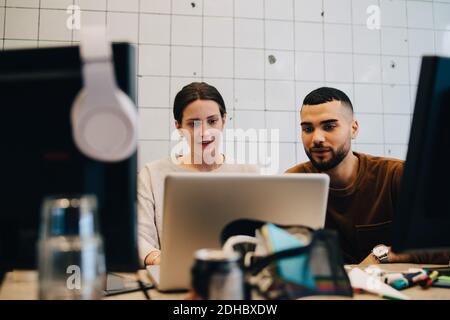 Confident young male and female colleagues looking at laptop against wall in office Stock Photo