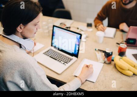 High angle view of female programmer writing on book by laptop while male colleague working at desk in creative office Stock Photo
