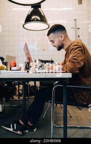 Full length side view of confident young male computer programmer writing while sitting with laptop at desk in small off Stock Photo