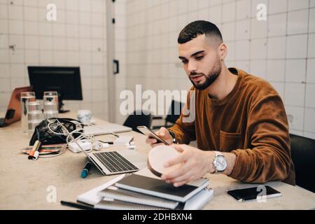 Young male hacker using smart phone while holding equipment at desk in small creative office Stock Photo