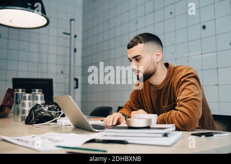 Confident young male computer programmer using laptop at desk in small office Stock Photo