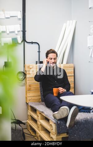 Creative businessman talking through mobile phone while sitting on stack of pallets in office Stock Photo