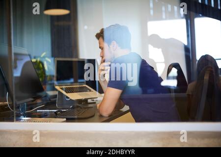 Businessman sitting at computer desk seen through glass in office Stock Photo