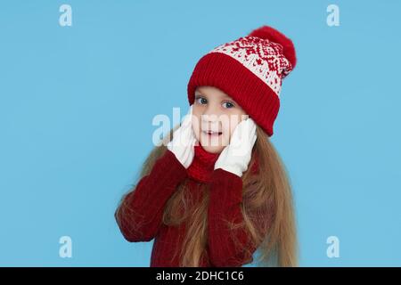 Winter portrait of happy child girl wearing knitted red hat, snood and sweater. Wow face. Blue background. childrens winter entertainment Stock Photo