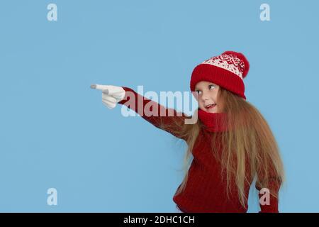 Winter portrait of happy child girl wearing knitted red hat, snood and sweater. shows her hand to the left. Blue background. childrens winter entertai Stock Photo