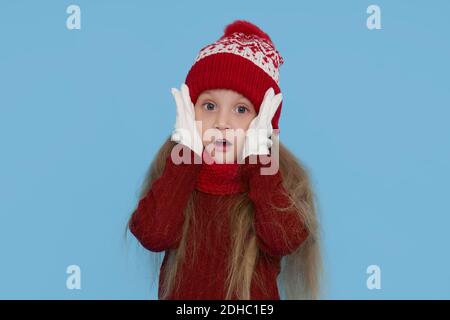 Winter portrait of happy child girl wearing knitted red hat, snood and sweater. Wow face. Blue background. childrens winter entertainment Stock Photo