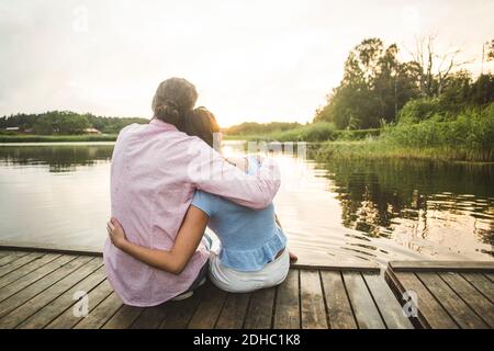 Rear view of couple sitting with arms around on jetty over lake during sunset Stock Photo