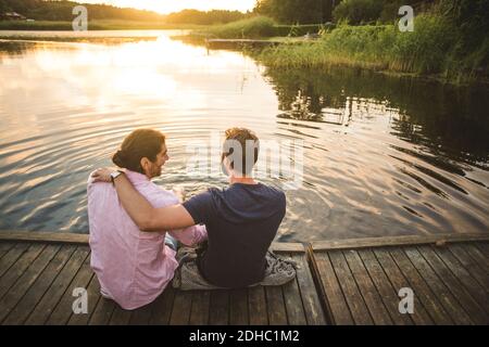 Rear view of smiling male friends sitting on jetty over lake during weekend Stock Photo