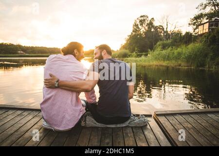 Rear view of smiling male friends sitting on jetty over lake during sunset Stock Photo