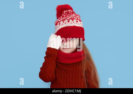 Winter portrait of happy child girl pulled a knitted red hat over her eyes, wearing snood and sweater. Wow face. Blue background. childrens winter ent Stock Photo