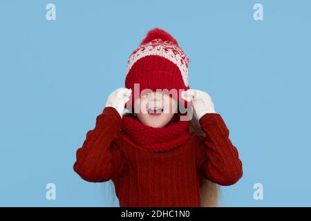 Winter portrait of happy child girl pulled a knitted red hat over her eyes, wearing snood and sweater. Wow face. Blue background. childrens winter ent Stock Photo