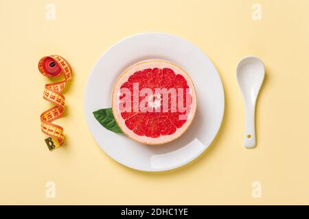 Halved ripe juicy grapefruit on a white plate, white spoon and measuring tape on a pastel yellow background. Slimming diet, body weight control. Stock Photo