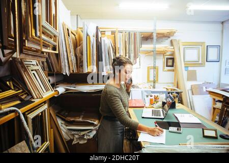 Confident young craftswoman talking on smart phone while writing in paper at workbench in framing store Stock Photo