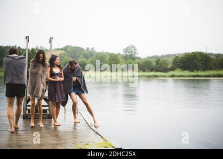 Male and female friends wrapped in towels walking on jetty over lake against clear sky Stock Photo