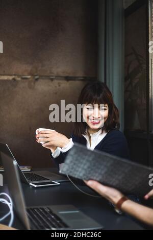 Smiling female entrepreneur holding coffee cup discussing with colleague over document at desk in office Stock Photo