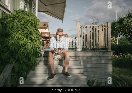 Full length of senior man using mobile phone while sitting on steps at porch Stock Photo