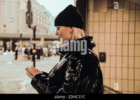 Side view of young woman using mobile phone while wearing black shiny jacket at subway's entrance in city during winter Stock Photo