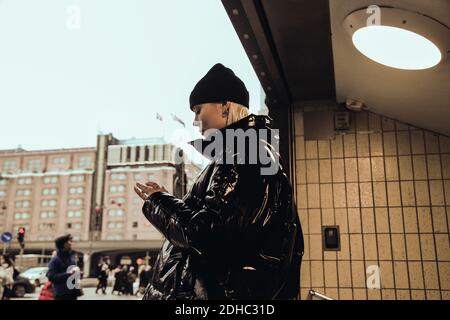 Side view of young woman wearing black shiny jacket while standing at subway's entrance in city during winter Stock Photo
