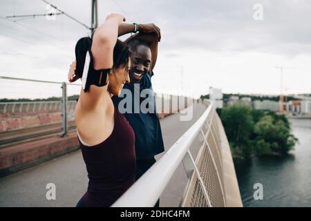 Smiling male and female athletes stretching hands on footbridge over sea against sky Stock Photo