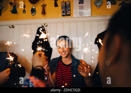 Smiling young multi-ethnic friends holding burning sparklers while enjoying in restaurant during dinner party Stock Photo