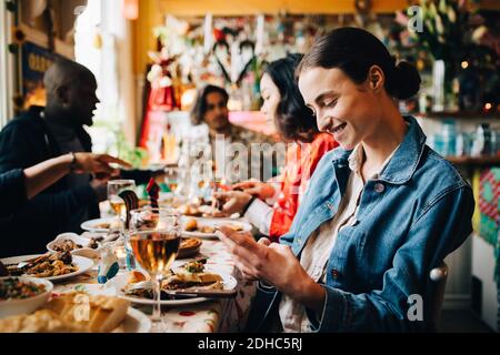 Smiling young woman using smart phone while sitting with friends at table in restaurant during dinner party Stock Photo