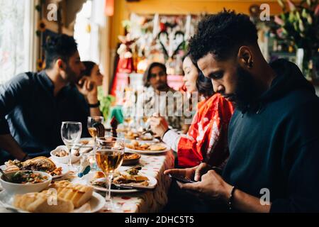 Young man text messaging through smart phone while sitting with friends during brunch party at restaurant Stock Photo
