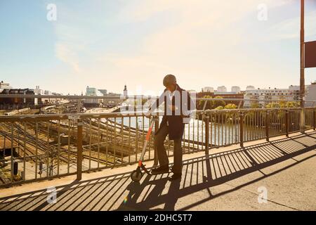 Full length of young man standing with electric push scooter by railing on bridge in city Stock Photo